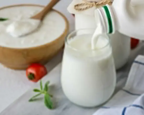 Ayran co to? Orzeźwiający napój mleczny, idealny na upalne dni, nalewany do szklanki. Obok miska z jogurtem i pomidorek.