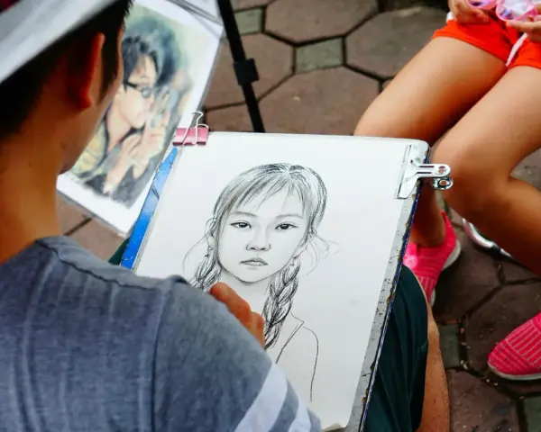 Artysta tworzy portret dziewczynki z warkoczami. Obok leży gotowy rysunek chłopca w okularach.