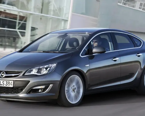 Ciemnoszary Opel Astra J 1.6 benzyna w ruchu, z kierowcą w okularach za kierownicą, na tle nowoczesnego budynku.