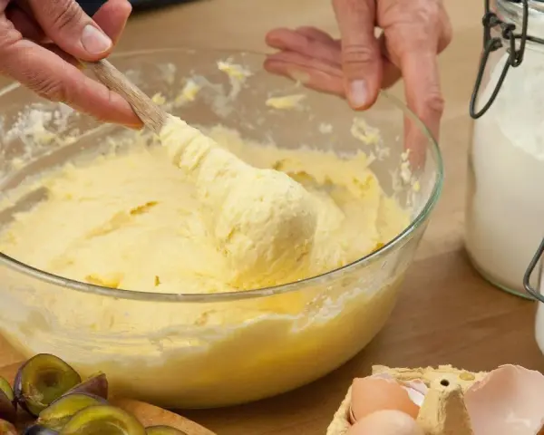 Hände rühren Teig mit Butter oder Margarine in einer Schüssel. Daneben liegen Pflaumen, Eier und Zucker.