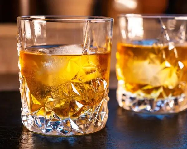 Zwei Gläser mit bernsteinfarbenem on the rocks drink, beleuchtet von warmem Licht.