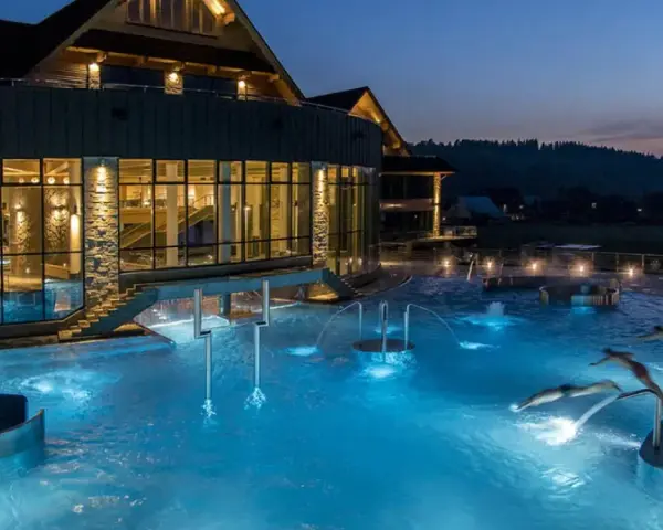 Nocą w Termach Chochołowskich hotel lśni światłem, a baseny zewnętrzne kuszą błękitem. Ludzie pływają i relaksują się w wodzie.