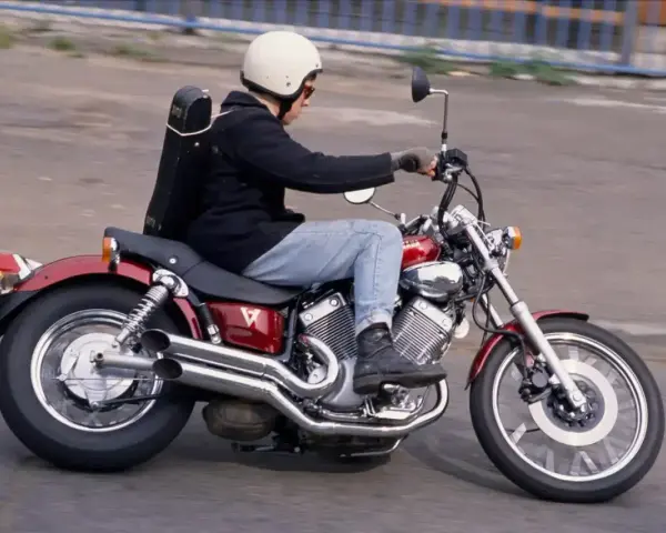Mężczyzna w kasku jedzie na czerwonym motocyklu Yamaha Virago 250 z futerałem na plecach.