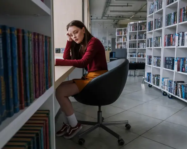 Zamyślona nastolatka w bibliotece, otoczona książkami, odzwierciedla złożoność zachowań nastolatków w okresie dojrzewania.