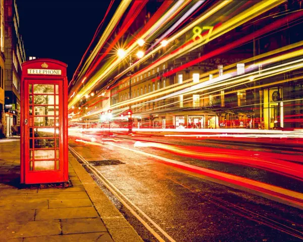 Klasyczna czerwona budka telefoniczna na tle nocnej ulicy, gdzie długi czas naswietlania zdjecia uchwycił smugi świateł samochodowych.
