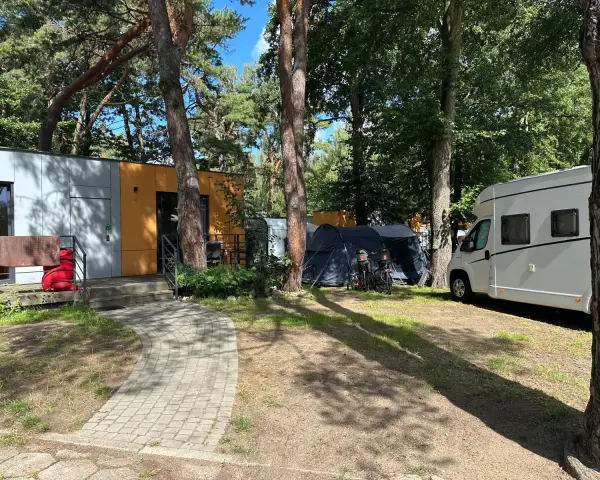 Na kempingu 45, wśród drzew, stoją nowoczesne domki, namioty i biała kamper.