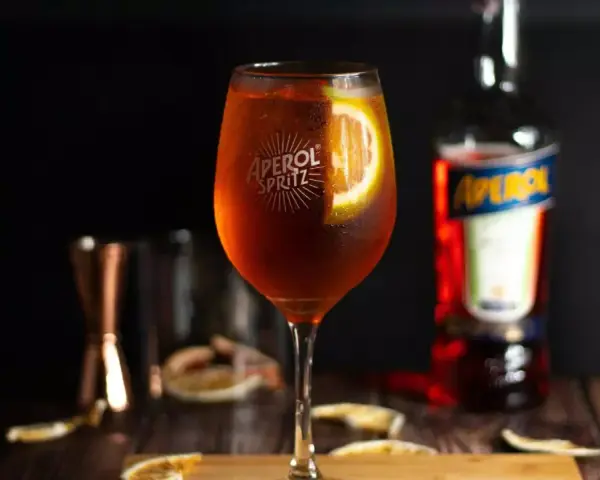 Orzeźwiający Aperol Spritz z plasterkiem pomarańczy, obok butelka Aperol. Idealny wybór zamiast Hugo.