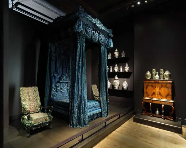 Cama con dosel azul, sillas ornamentadas y vitrinas con cerámica, como si fueran escenarios de cursos de arte online.
