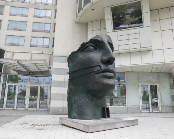 Okaleczona, ciemna rzeźba głowy z opaską na ustach, na tle nowoczesnego budynku.