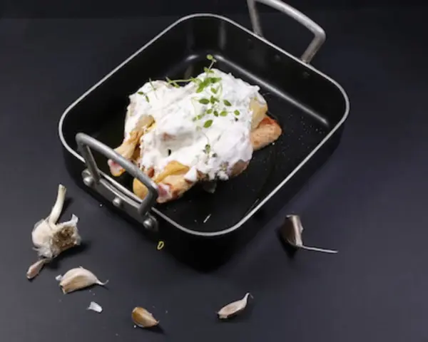 Un poulet entier est nappé de fromage blanc crémeux et garni d'herbes fraîches dans un plat noir. Des gousses d'ail sont disposées autour.