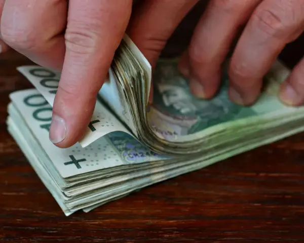 Dłonie przewracają stos banknotów 100 zł na drewnianym blacie.