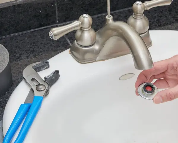Hand holding a removed faucet aerator with red buildup, next to pliers and duct tape on a sink.