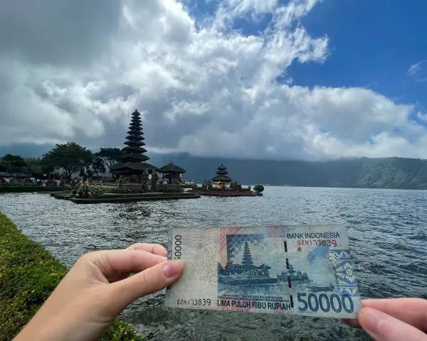 Ręka trzyma banknot 50 000 rupii na tle świątyni na wodzie. To symboliczne, że wiza turystyczna na Bali jest w zasięgu ręki!