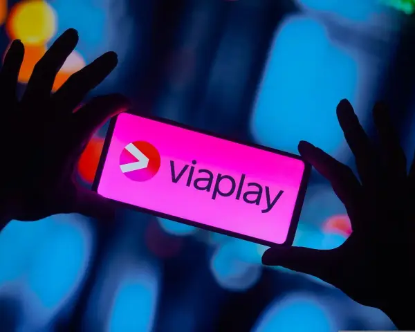 Dłonie trzymają smartfon z logo Viaplay. W tle rozmyte, kolorowe światła.