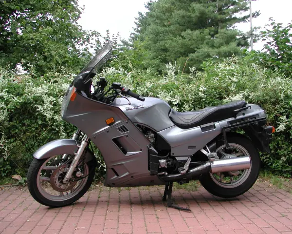 Srebrny motocykl Kawasaki GTR 1000 zaparkowany na czerwonej kostce brukowej, otoczony zielenią.