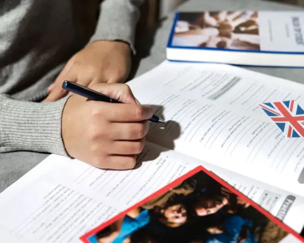 Osoba pisze długopisem w zeszycie z ćwiczeniami z języka angielskiego. Obok leży książka i flaga Wielkiej Brytanii. To może być przygotowanie do napisania klauzuli CV po angielsku.