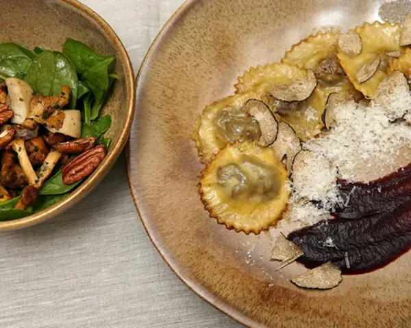 Ravioli pasta mit Trüffeln, Parmesan und einer dunklen Sauce. Daneben eine Schüssel mit Pilzen, Nüssen und Spinat.