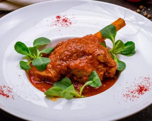 Pierna de cordero en salsa roja con hojas verdes en plato blanco.