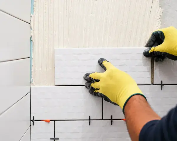 Mains gantées de jaune en train de coller du carrelage sur du carrelage, avec des croisillons pour un espacement parfait.