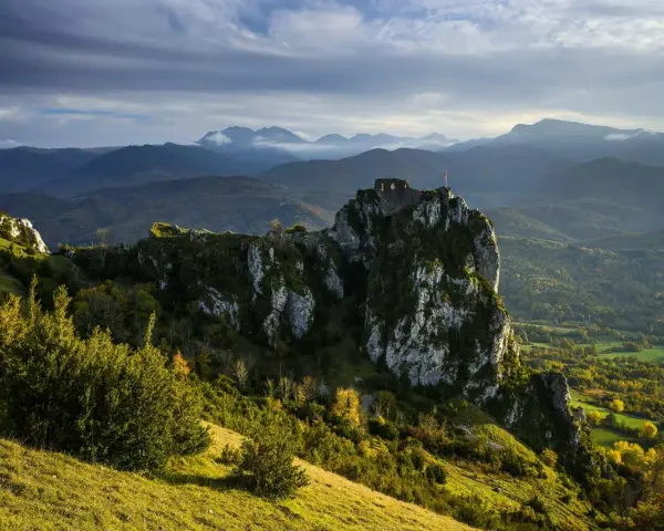 Ruines d'un château cathare sur un pic rocheux, dominant un paysage de montagnes et de forêts. Le chemin des bonshommes serpente à travers la vallée.