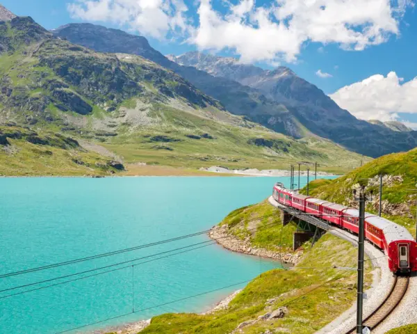 A red train winds along a track beside a vibrant turquoise lake, with lush green mountains and a bright blue sky in the background. This is a classic como to switzerland train journey.