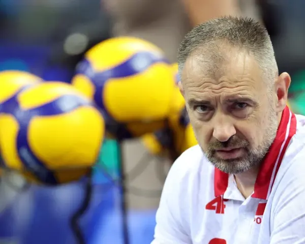 Skupiony Nikola Grbić, trener siatkówki, w białej koszulce z czerwonymi akcentami, na tle żółto-niebieskich piłek.