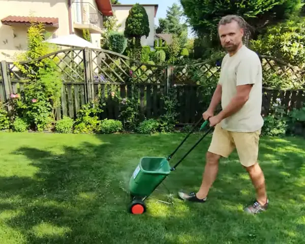 Mężczyzna z dredami pokazuje, jak ustawić siewnik do trawy, rozsypując nawóz na trawniku.