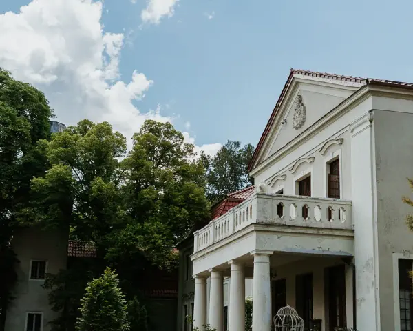 Pałac Grąbkowo, biały budynek z kolumnami i balkonem, otoczony bujną zielenią drzew pod błękitnym niebem z chmurami.