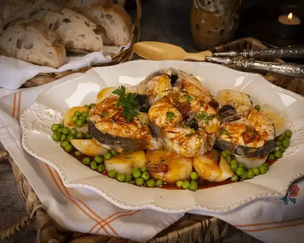 Merluza a la gallega servida en plato blanco con patatas, guisantes y perejil. Pan rústico en cesta al fondo.