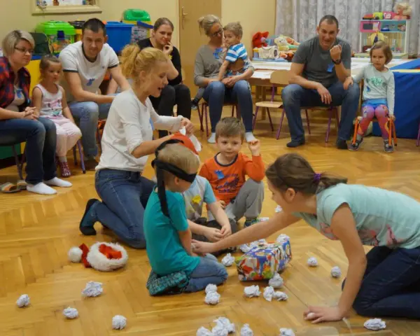Zajęcia integracyjne z rodzicami w przedszkolu: dzieci z zawiązanymi oczami szukają prezentu, rodzice obserwują.