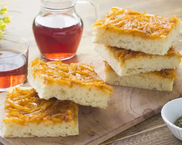 Focaccia con cipolle dorate e soffice, servita su un tagliere di legno con un bicchiere di vino rosato.