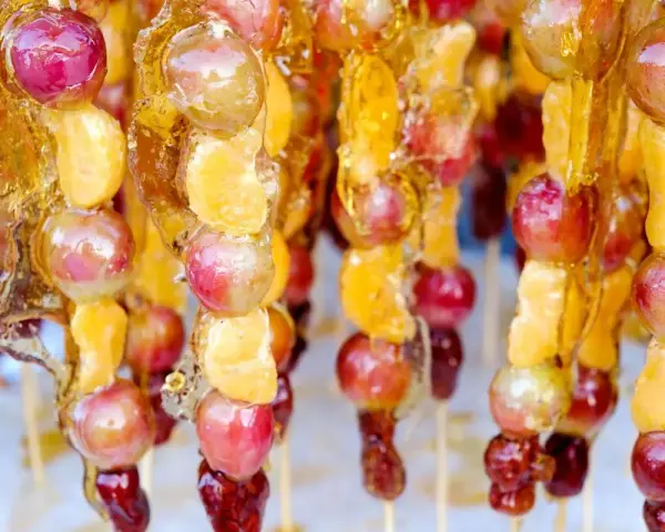Spiedini di frutta caramellata con acqua e zucchero, con uva e spicchi d'arancia lucidi e dorati.