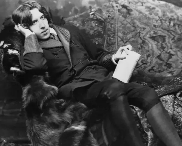 Oscar Wilde, w futrzanym płaszczu, z książką w dłoni, w pozie sugerującej refleksję nad swoimi sztukami.