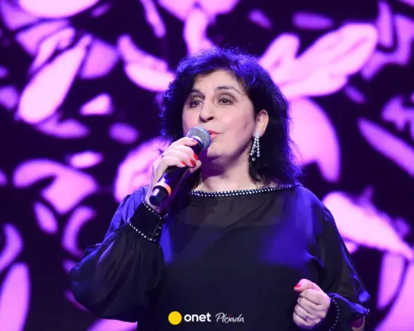 Kobieta śpiewa na scenie, w czarnej, ozdobnej sukni. Wiek męża Eleni nie jest widoczny na zdjęciu.
