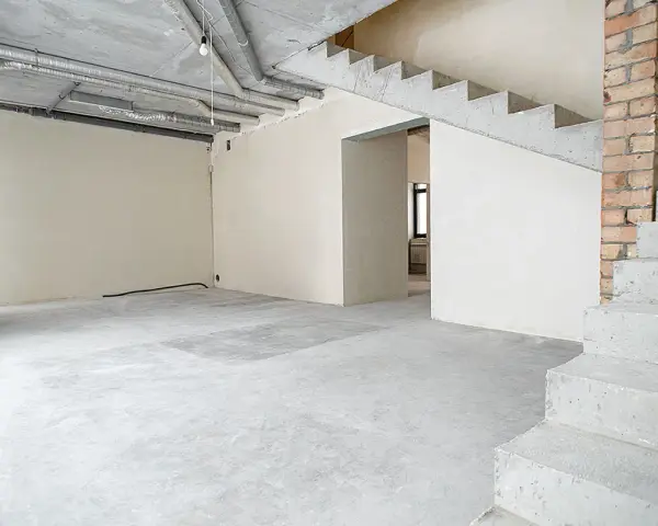 Wnętrze w stanie deweloperskim: betonowe schody, nieotynkowane ściany, widoczne instalacje i przewody.
