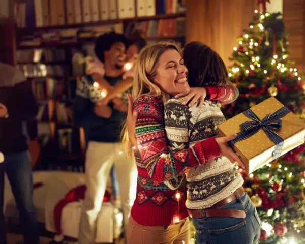 Zwei Frauen umarmen sich vor einem Weihnachtsbaum, eine hält ein Geschenk. Eine Familie feiert Weihnachten, eine der vielen schönen weihnachtstraditionen deutschland.