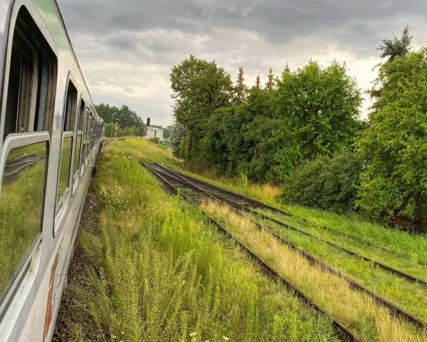 Widok z okna pociągu na tory kolejowe otoczone zielenią i chmury na niebie.