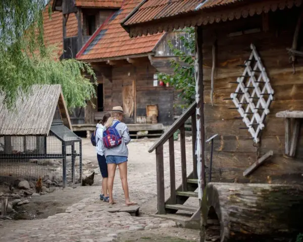 Dwie dziewczyny zwiedzają skansen, który może być częścią oferty hotelu bon turystyczny. W tle drewniane chaty i kurnik.