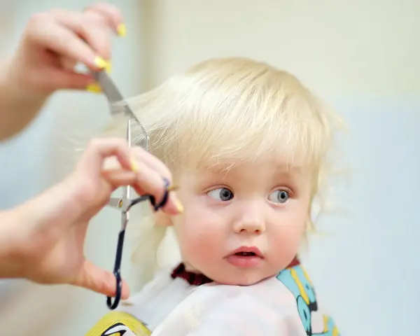 Ein kleines blondes Kind bekommt die Haare geschnitten. Die Schere nähert sich vorsichtig den feinen Strähnen.
