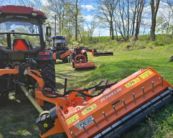 Pomarańczowy mulczer używany, podłączony do traktora, gotowy do pracy na zielonej łące.