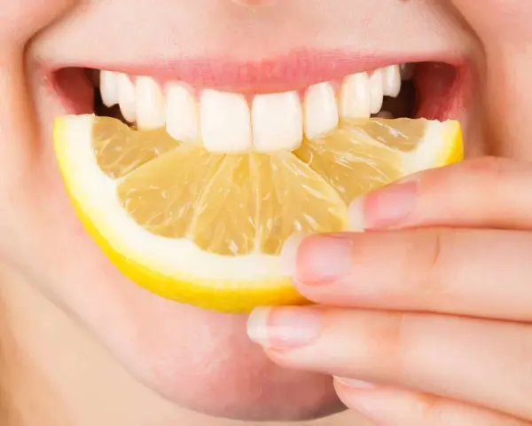 Une personne tient une tranche de citron près de sa bouche ouverte, révélant des dents blanches. Le citron est connu pour ses bienfaits sur la peau.
