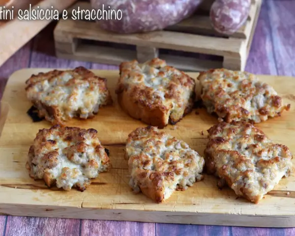 Sei crostini salsiccia e stracchino dorati e filanti, pronti per essere gustati su un tagliere di legno.