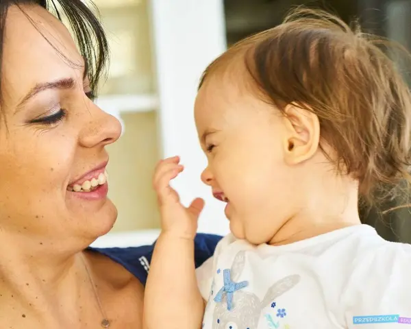 Uśmiechnięta mama patrzy na swoje dziecko, które bawi się i śmieje, wyciągając rączkę.