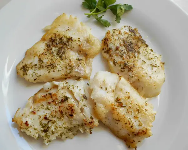 Cuatro trozos de pescado blanco rosada a la plancha, sazonados con hierbas, servidos en un plato blanco con una ramita de perejil.
