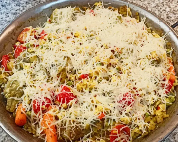 Arroz a la milanesa listo para hornear, cubierto con queso rallado y vegetales frescos como tomates, zanahorias y guisantes.
