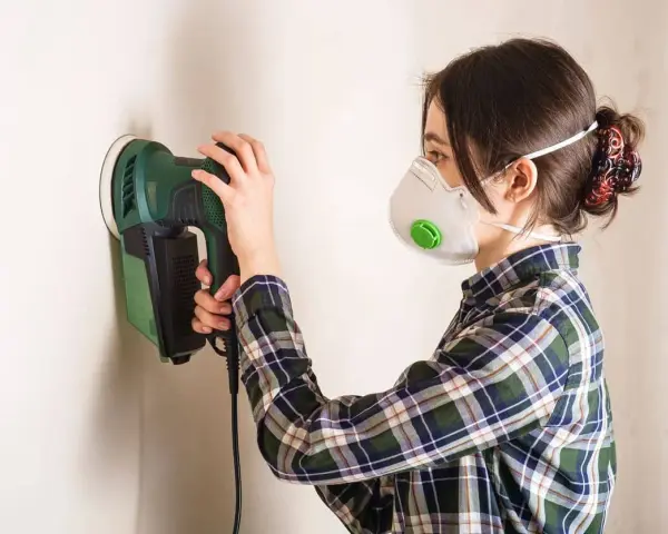 Persona con mascarilla y camisa a cuadros usa una lijadora eléctrica para lijar una pared.