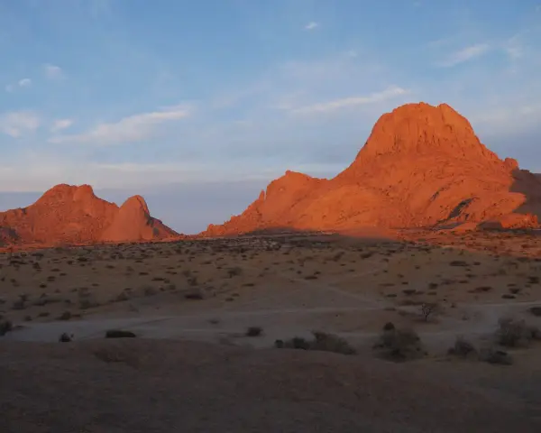 Zachód słońca maluje na pomarańczowo skalne szczyty Spitzkoppe w Namibii. Pustynny krajobraz z suchymi krzewami i ścieżkami.