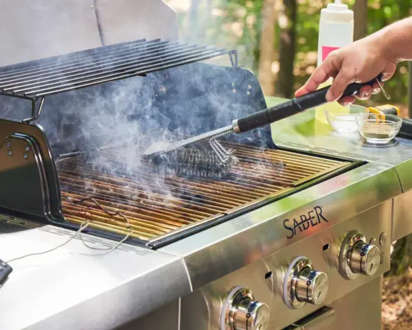 A hand uses a grill brush to clean the grates of a smoking Saber grill, showing the best way to clean a grill for your next cookout.