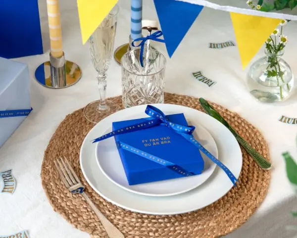 Blå och gula girlanger, ljus och presenter på ett dukat bord. En blå present med band och texten "Fy fan vad du är bra" är en fin pom pom dekoration.