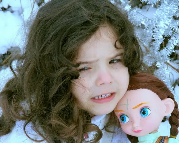 Ein Kind mit dunklen Haaren hält eine Puppe im Schnee. Das Kind sieht besorgt aus, als ob mein Kind Angst vor mir hätte.
